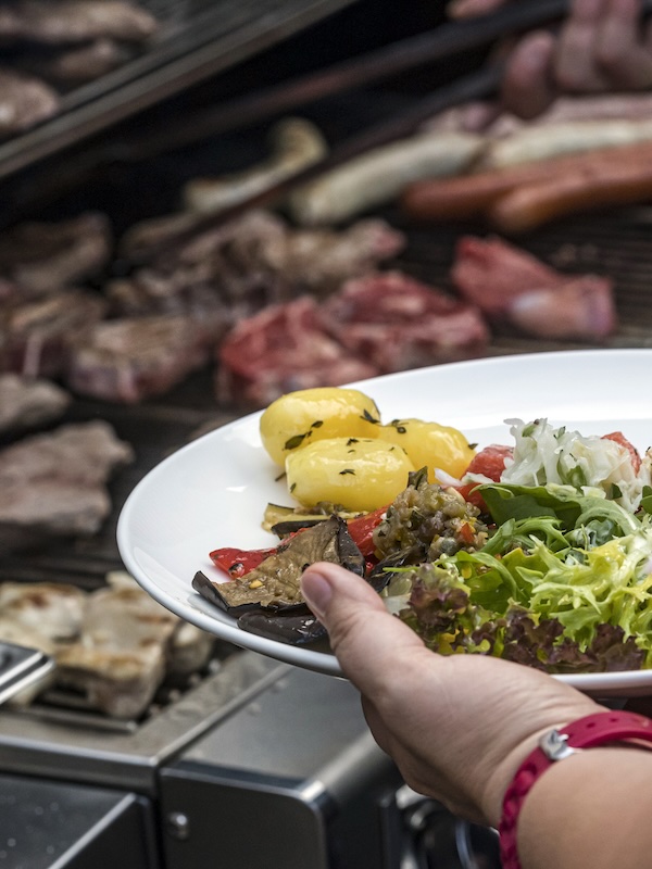 Weststadt Bar Catering, Person hält Teller mit Salat und Kartoffeln in der Hand, im Hintergrund sieht man einen Grill mit Fleisch
